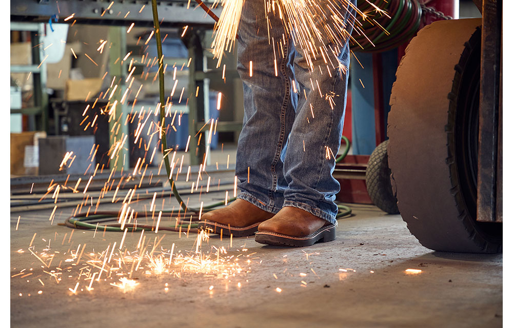 Las chispas saltan cuando Wes Stevenson, en jeans y botas de trabajo de cuero marrón, opera una herramienta de corte en un taller de metal, con el enfoque en sus piernas y pies.